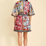 A woman viewed from the back wearing a short, collared kimono-style jacket with a colorful paisley and floral print in red, blue, yellow, and white, paired with black high heels against a solid beige background.