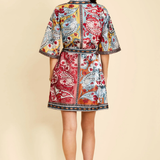 A woman viewed from the back wearing a short, collared kimono-style jacket with a colorful paisley and floral print in red, blue, yellow, and white, paired with black high heels against a solid beige background.