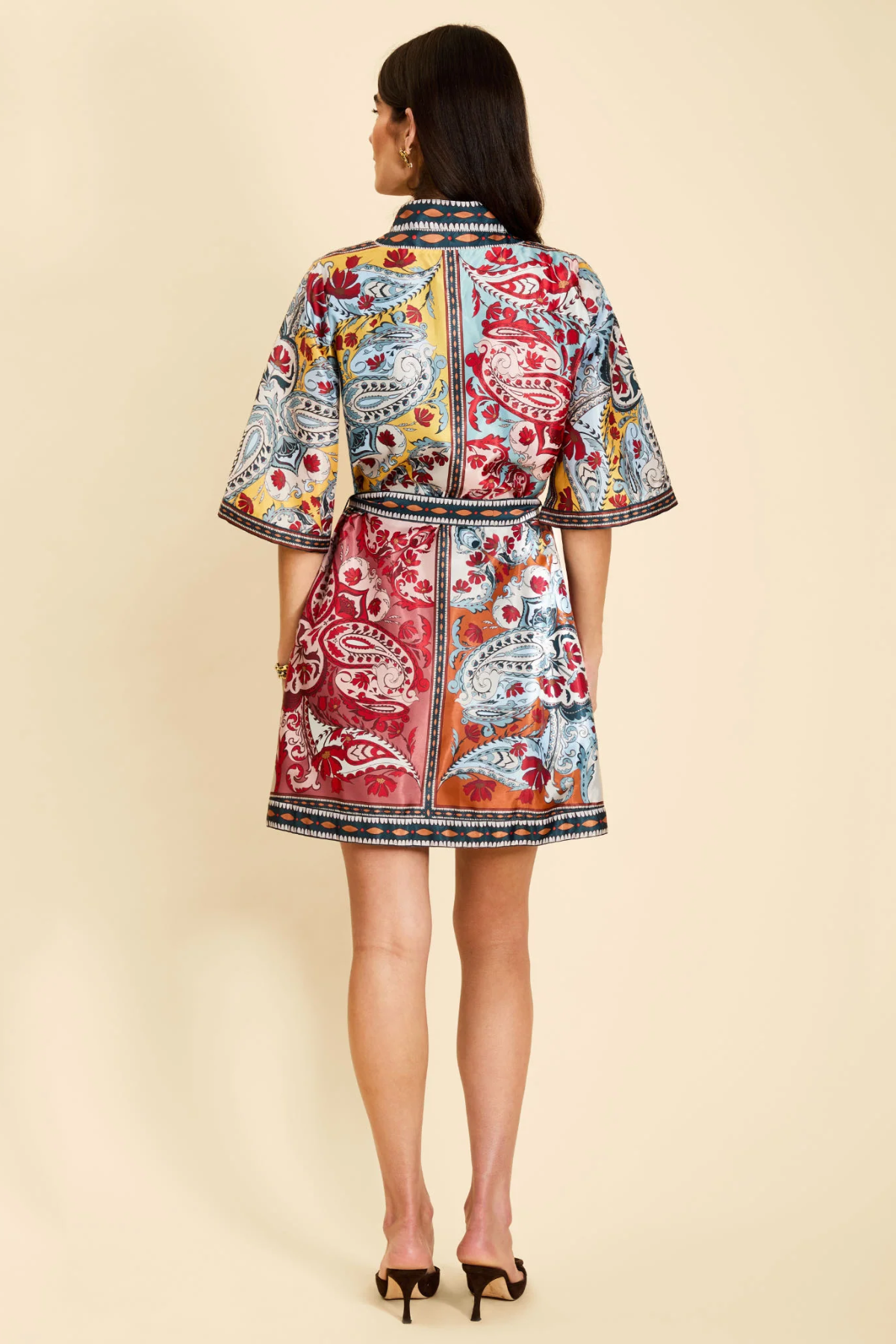 A woman viewed from the back wearing a short, collared kimono-style jacket with a colorful paisley and floral print in red, blue, yellow, and white, paired with black high heels against a solid beige background.