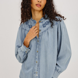 A woman with curly dark hair wearing a light blue denim button-up shirt with embroidered details on the shoulders and chest, paired with blue jeans, against a plain light gray studio background.