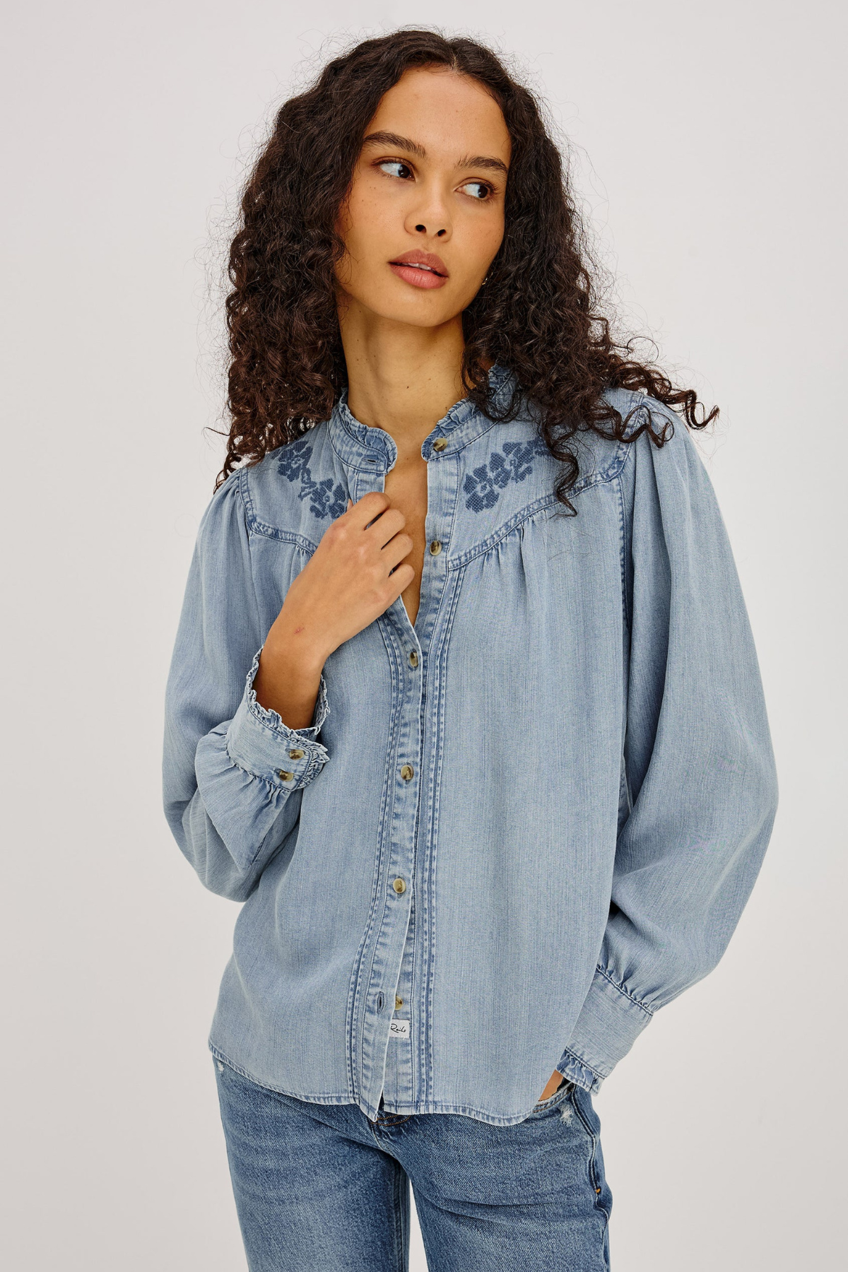 A woman with curly dark hair wearing a light blue denim button-up shirt with embroidered details on the shoulders and chest, paired with blue jeans, against a plain light gray studio background.