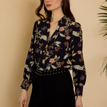 A woman modeling a long-sleeve floral print blouse with a deep V-neck and button-front closure, paired with black pants, against a warm beige background with a plant leaf visible on the right.