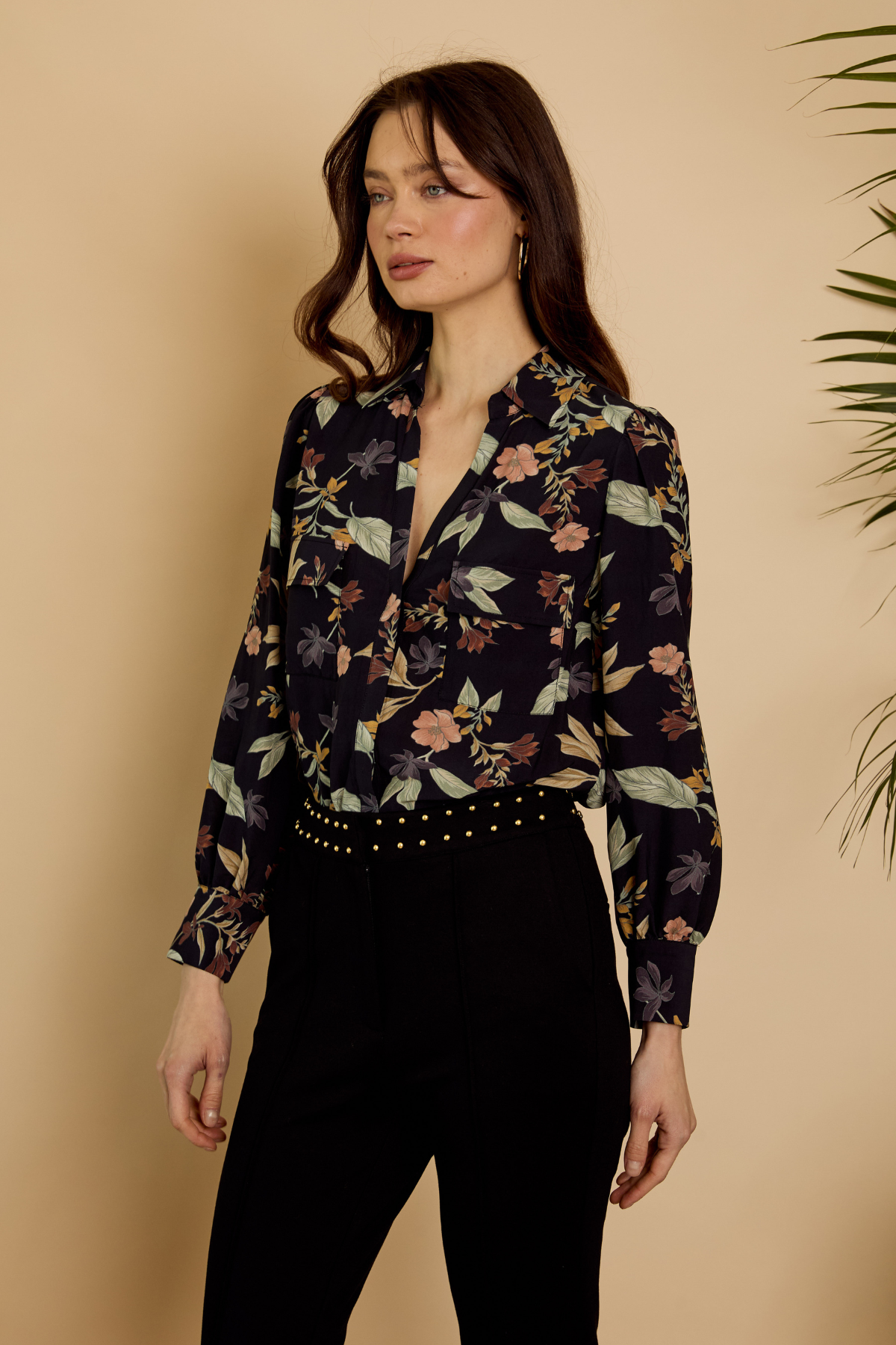 A woman modeling a long-sleeve floral print blouse with a deep V-neck and button-front closure, paired with black pants, against a warm beige background with a plant leaf visible on the right.