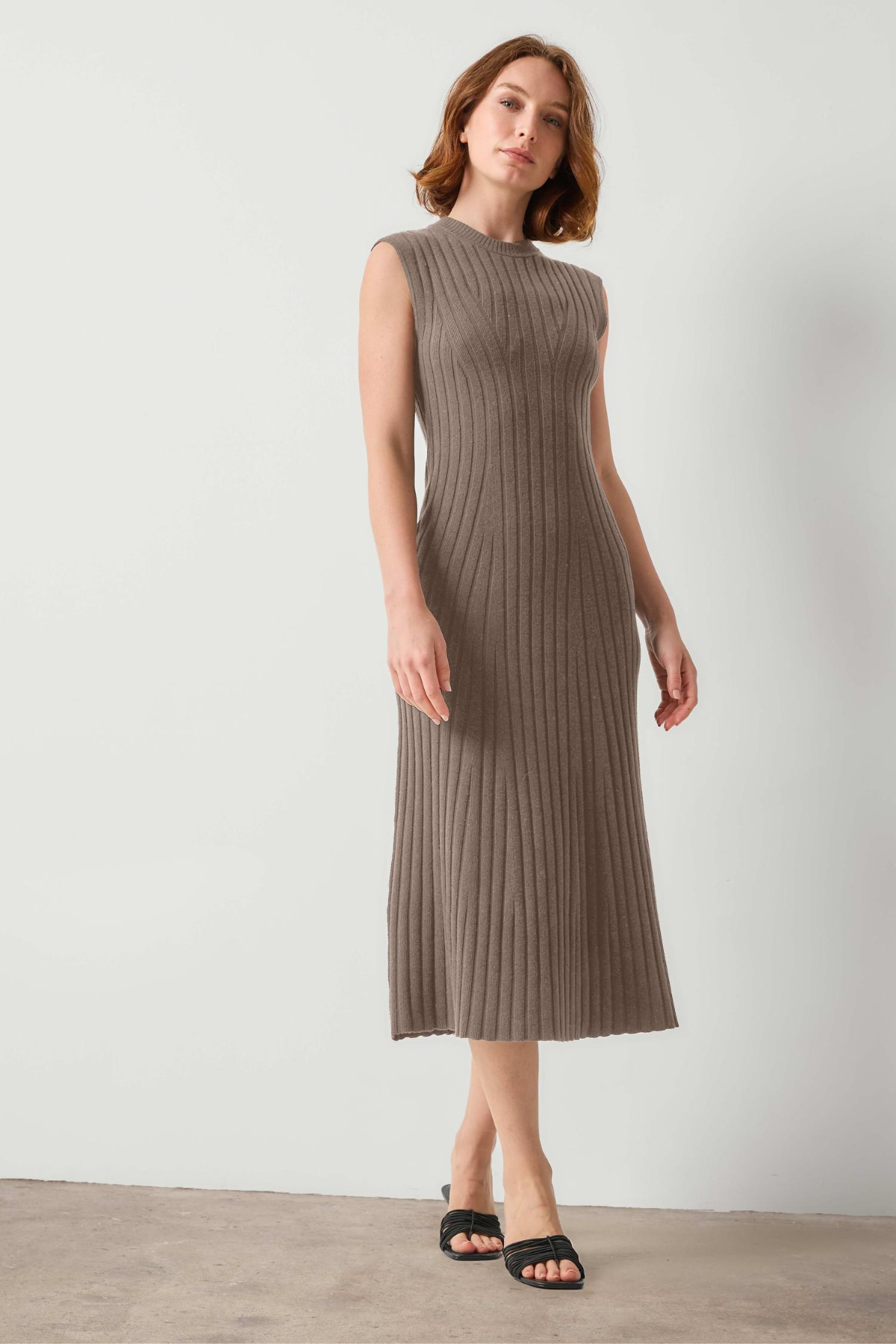A woman modeling a sleeveless, ribbed knit midi dress in a taupe-brown color, paired with black slide sandals, against a plain light gray studio background.