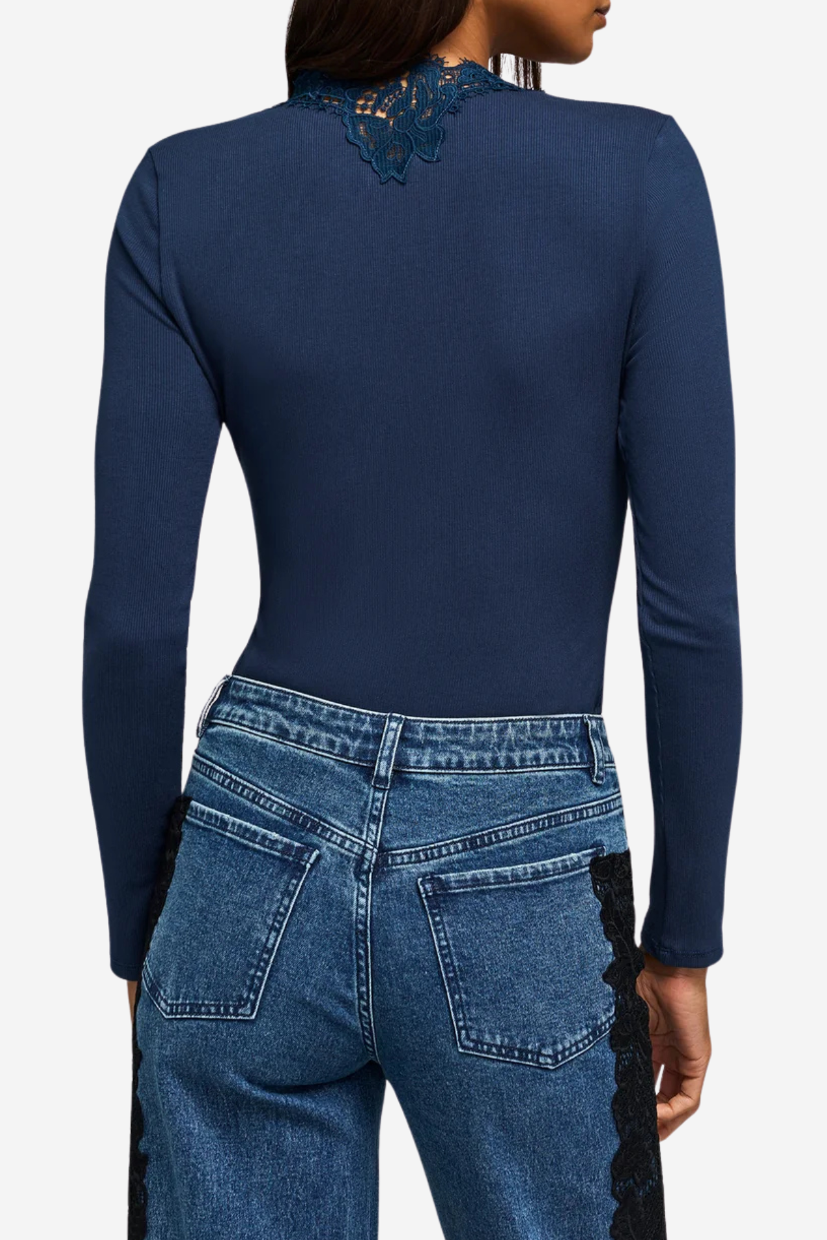 A woman shown from the back wearing a navy blue long-sleeve top with lace detailing at the neckline and blue jeans with black lace side panels, against a light gray studio background.