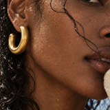 A close-up lifestyle shot of a woman wearing a thick, polished gold huggie hoop earring. Her face is in profile, with wet, glistening hair and a hint of blue sky in the background.