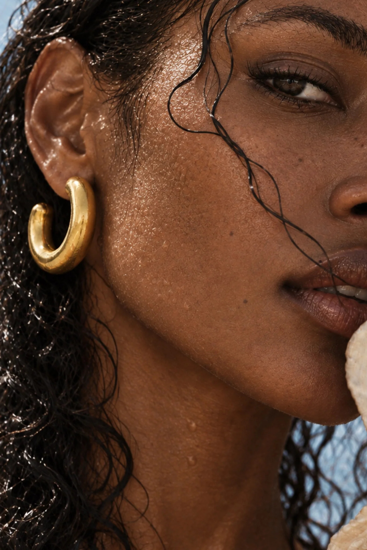A close-up lifestyle shot of a woman wearing a thick, polished gold huggie hoop earring. Her face is in profile, with wet, glistening hair and a hint of blue sky in the background.