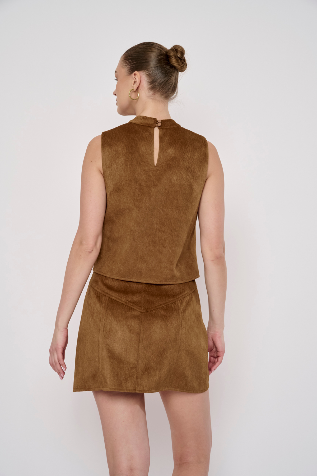 A woman shown from the back wearing a sleeveless brown faux-suede mini dress with a high keyhole neckline and a matching mini skirt, against a plain light gray studio background.