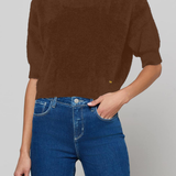 A woman wearing a brown short-sleeve knit sweater and blue jeans, cropped from the mouth down against a light gray studio background.