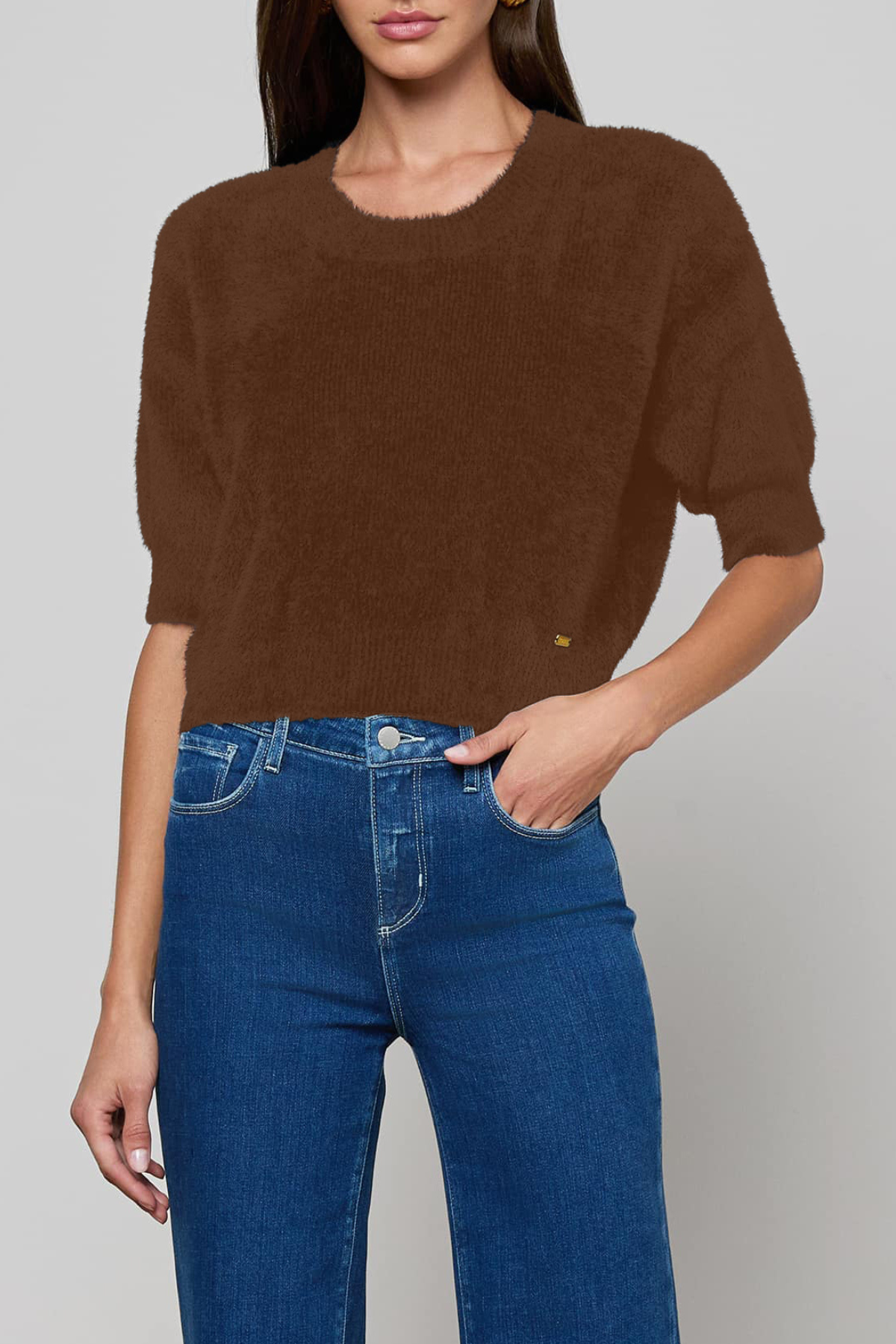 A woman wearing a brown short-sleeve knit sweater and blue jeans, cropped from the mouth down against a light gray studio background.