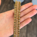 A hand holding a bracelet decorated with gold beads, set against a blurred gray background.