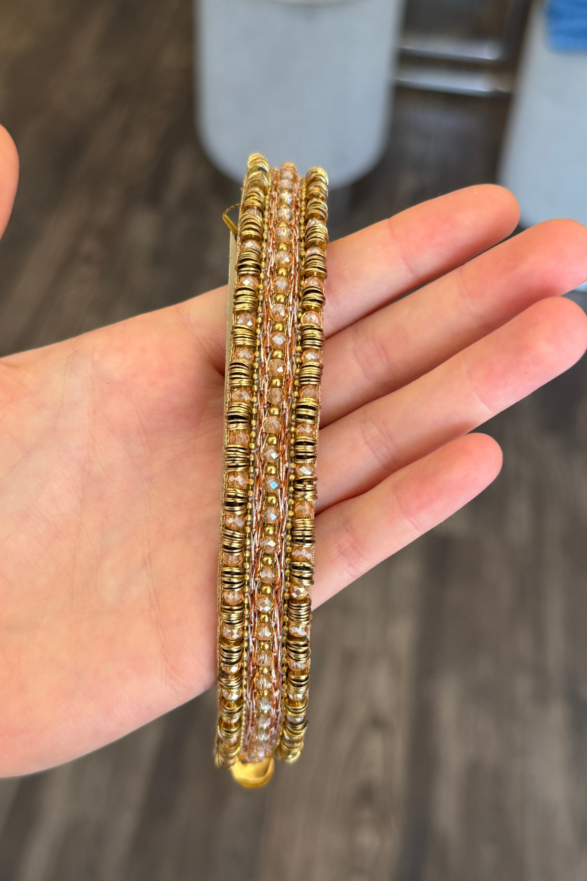 A hand holding a bracelet decorated with gold beads, set against a blurred gray background.