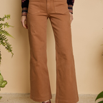 A person wearing tan wide-leg cropped pants with a gold button and belt, paired with a patterned long-sleeve top and brown heeled sandals, standing against a beige wall with plants.