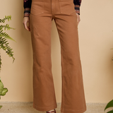A person wearing tan wide-leg cropped pants with a gold button and belt, paired with a patterned long-sleeve top and brown heeled sandals, standing against a beige wall with plants.