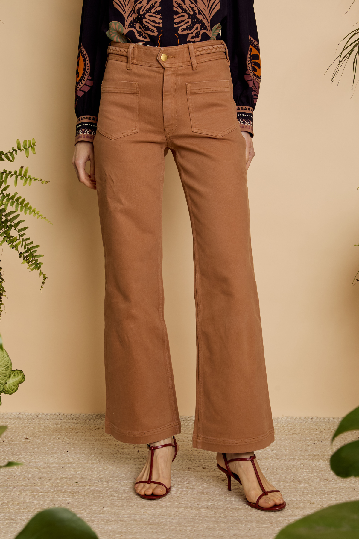 A person wearing tan wide-leg cropped pants with a gold button and belt, paired with a patterned long-sleeve top and brown heeled sandals, standing against a beige wall with plants.