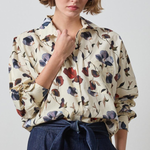 A woman wearing a cream-colored long-sleeve blouse with a blue, red, and brown floral print, paired with a dark blue high-waisted tie-front skirt, against a light gray studio background.
