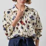 A woman wearing a cream-colored long-sleeve blouse with a blue, red, and brown floral print, paired with a dark blue high-waisted tie-front skirt, against a light gray studio background.