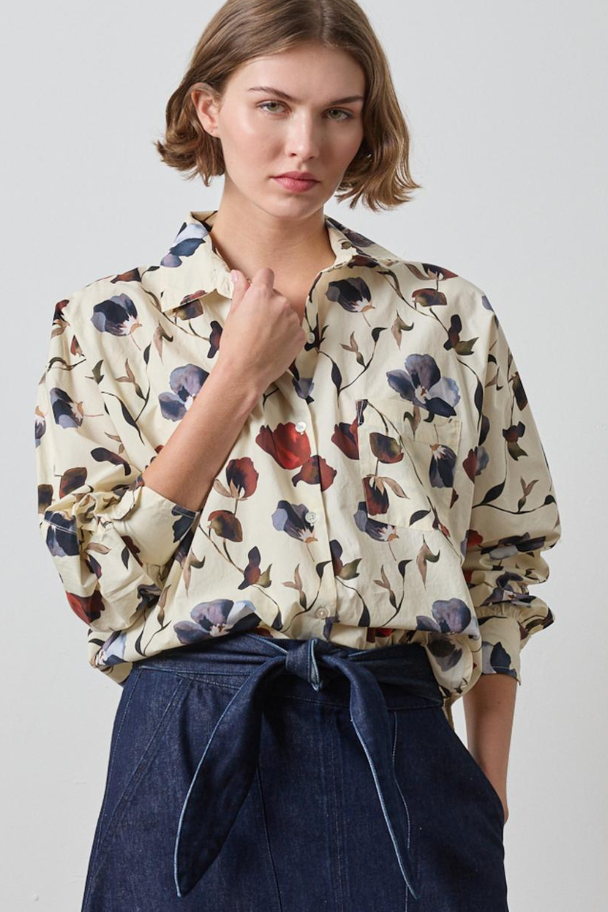 A woman wearing a cream-colored long-sleeve blouse with a blue, red, and brown floral print, paired with a dark blue high-waisted tie-front skirt, against a light gray studio background.