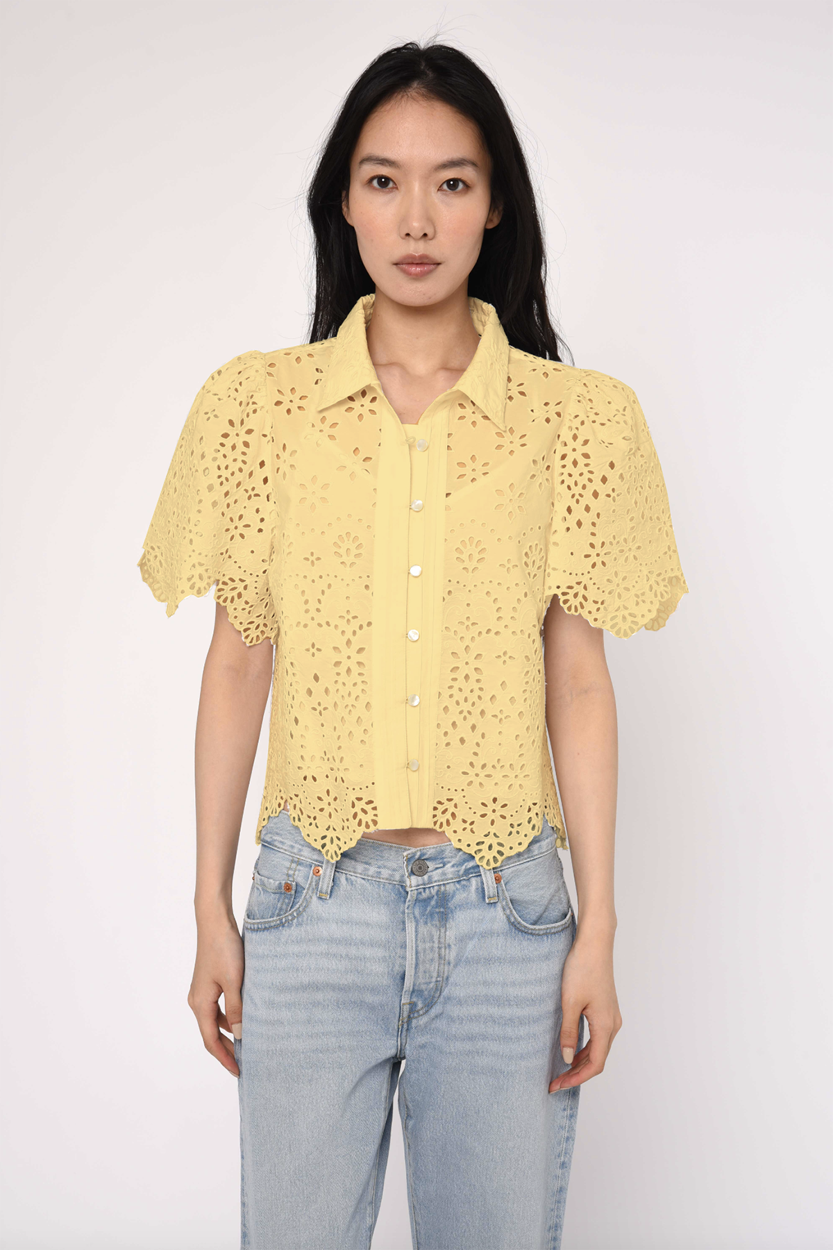 Woman wearing a yellow lace blouse and light blue jeans against a white background