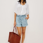Woman wearing a white blouse and denim shorts, holding a brown tote bag on a white background
