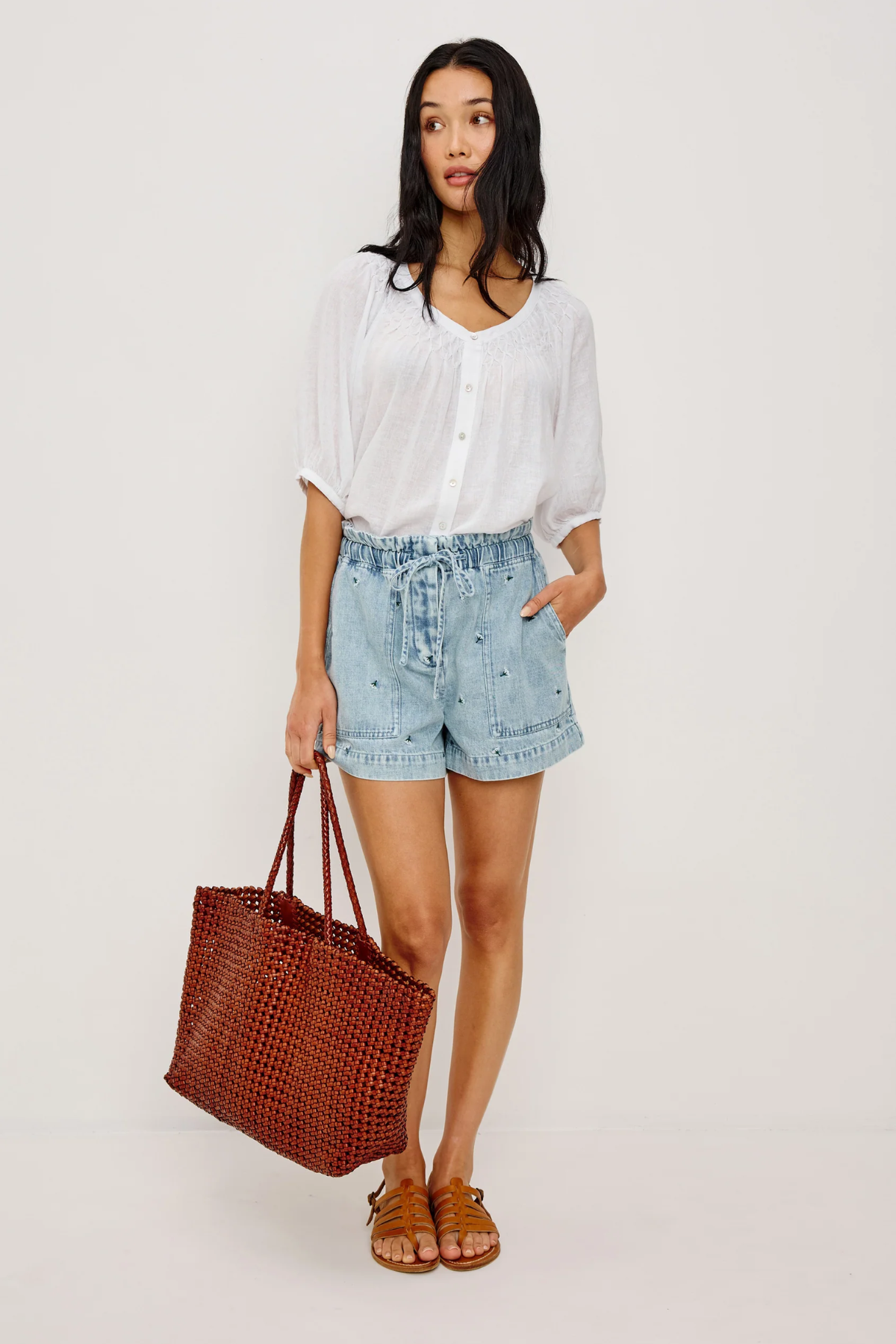 Woman wearing a white blouse and denim shorts, holding a brown tote bag on a white background