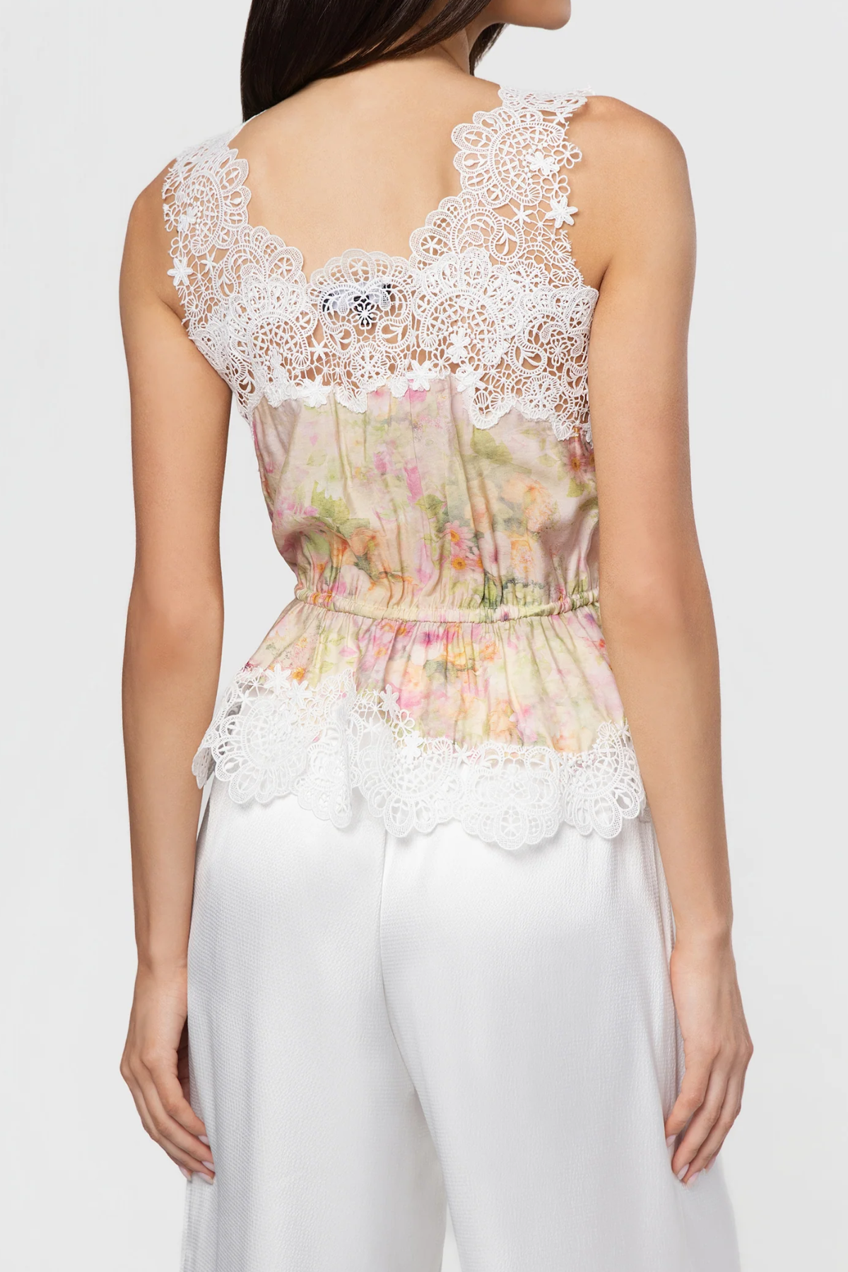 Woman wearing a floral top with lace details and white pants on a plain background
