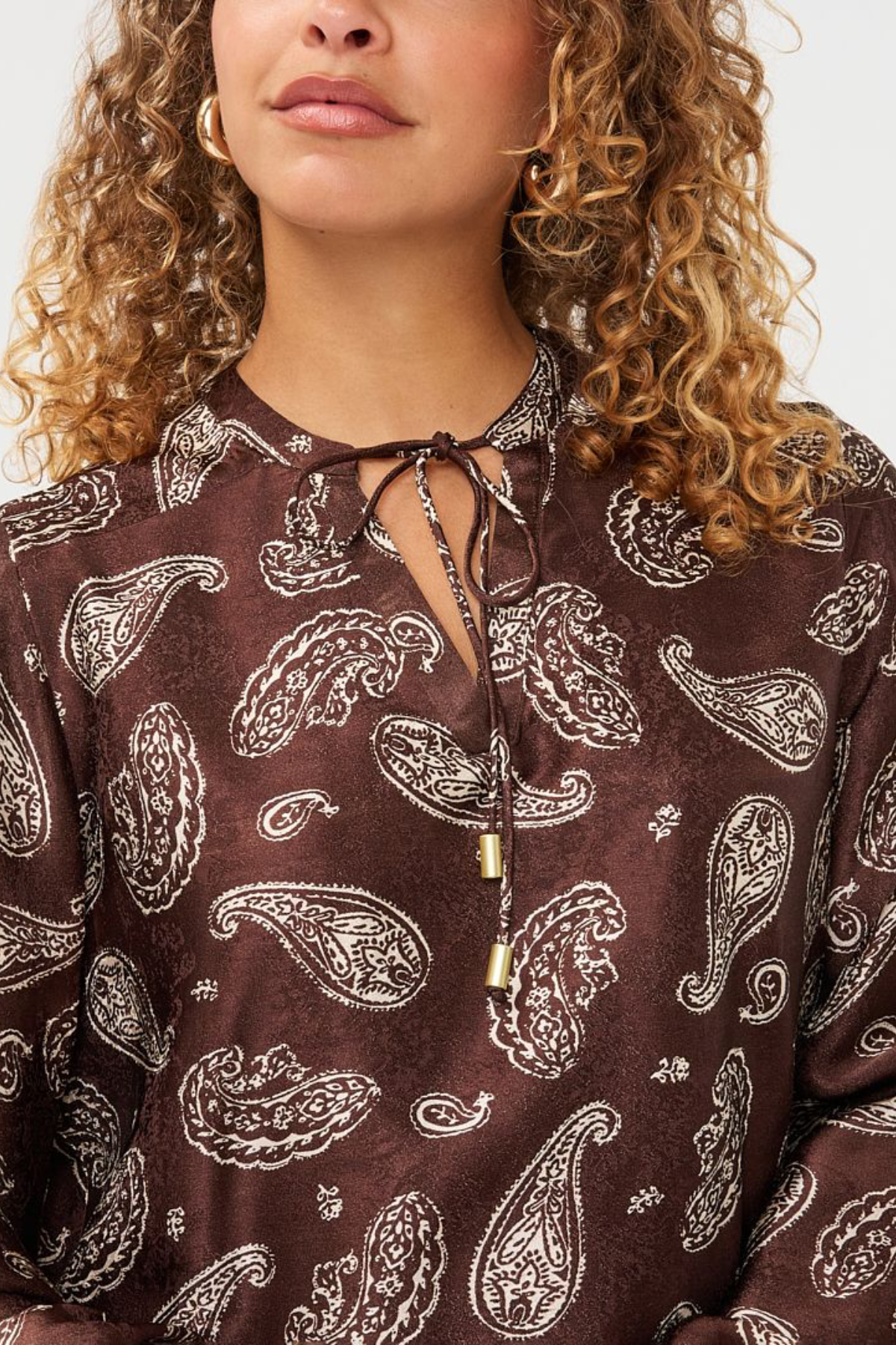 A close-up of a woman wearing a brown long-sleeve blouse with a white paisley pattern and a keyhole tie neckline.