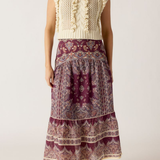 A woman modeling a sleeveless bohemian-style maxi dress featuring a cream knit ruffle bodice and a tiered purple paisley skirt, paired with tan ankle boots against a light beige studio background.