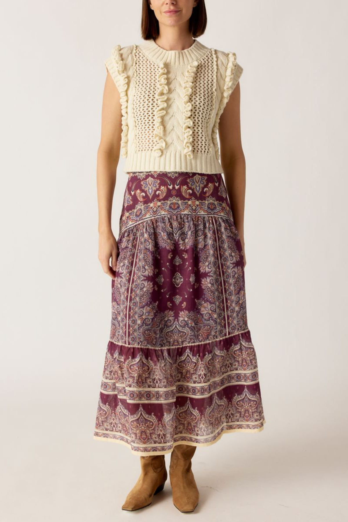 A woman modeling a sleeveless bohemian-style maxi dress featuring a cream knit ruffle bodice and a tiered purple paisley skirt, paired with tan ankle boots against a light beige studio background.