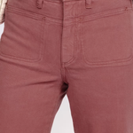 Close-up of a person wearing dusty rose or mauve-colored denim jeans, showing the front waistband, button, belt loops, and front patch pockets. A hand is resting near the waistband, and a black top is visible above the jeans.