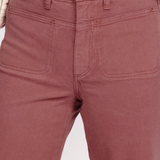 Close-up of a person wearing dusty rose or mauve-colored denim jeans, showing the front waistband, button, belt loops, and front patch pockets. A hand is resting near the waistband, and a black top is visible above the jeans.