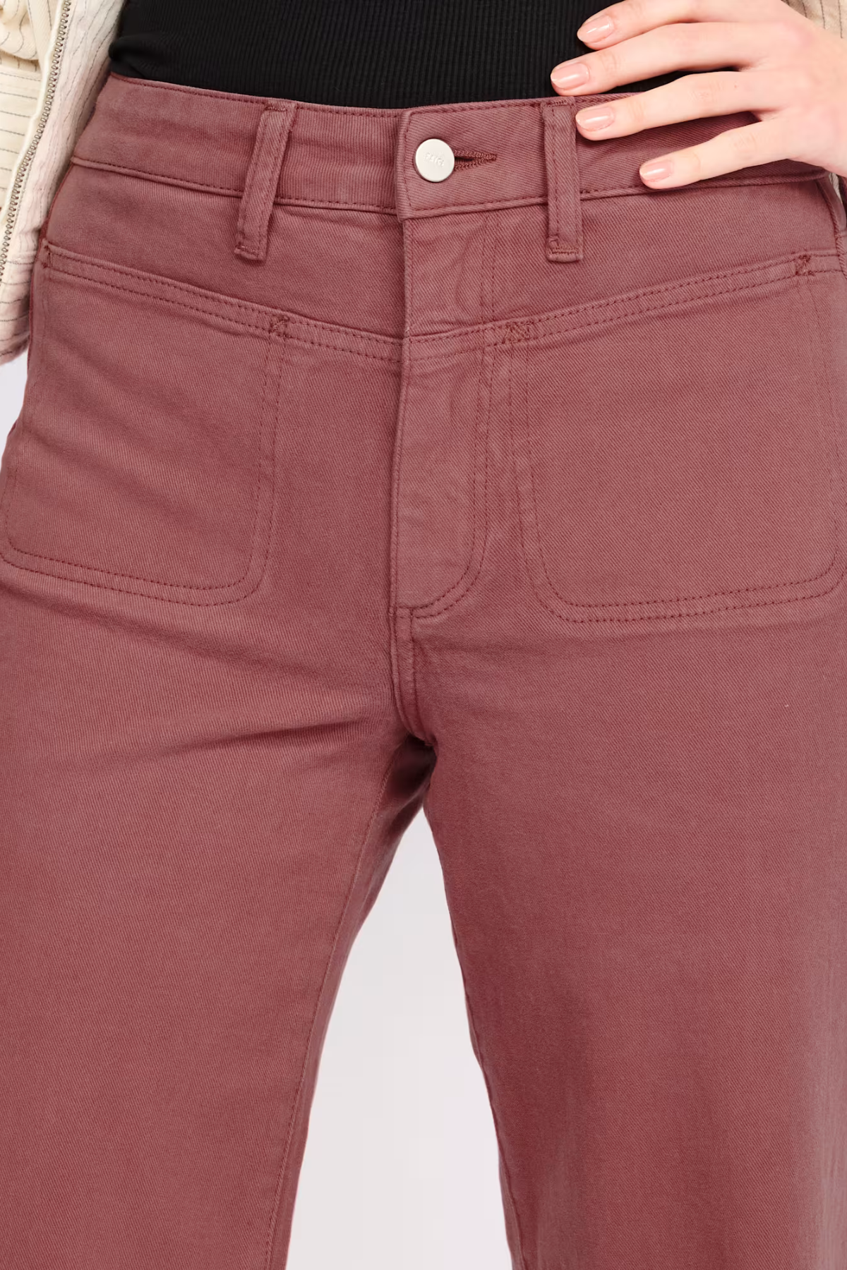 Close-up of a person wearing dusty rose or mauve-colored denim jeans, showing the front waistband, button, belt loops, and front patch pockets. A hand is resting near the waistband, and a black top is visible above the jeans.