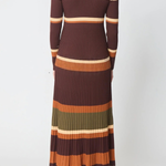 A woman viewed from the back wearing a long-sleeve ribbed knit maxi dress with horizontal stripes in dark brown, rust orange, cream, olive green, and brown, paired with brown heeled boots against a light gray studio background.