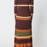 A woman viewed from the back wearing a long-sleeve ribbed knit maxi dress with horizontal stripes in dark brown, rust orange, cream, olive green, and brown, paired with brown heeled boots against a light gray studio background.