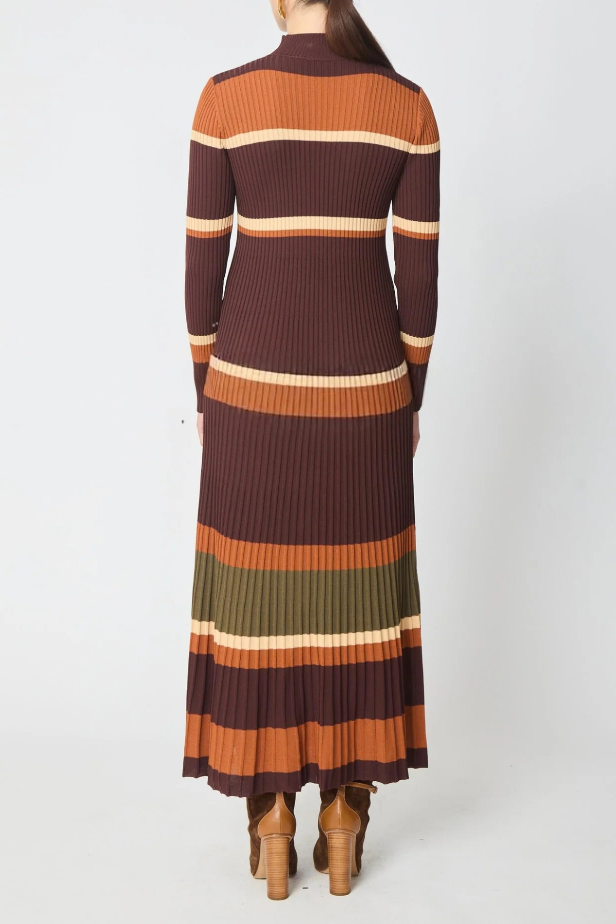 A woman viewed from the back wearing a long-sleeve ribbed knit maxi dress with horizontal stripes in dark brown, rust orange, cream, olive green, and brown, paired with brown heeled boots against a light gray studio background.