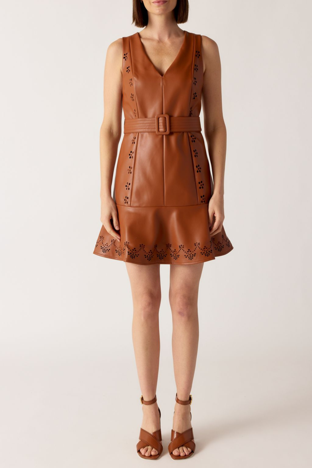A woman modeling a sleeveless brown faux-leather mini dress with a V-neck, matching belt, and floral embroidery on the bodice and flared hem, paired with brown strappy heeled sandals against a light beige studio background.