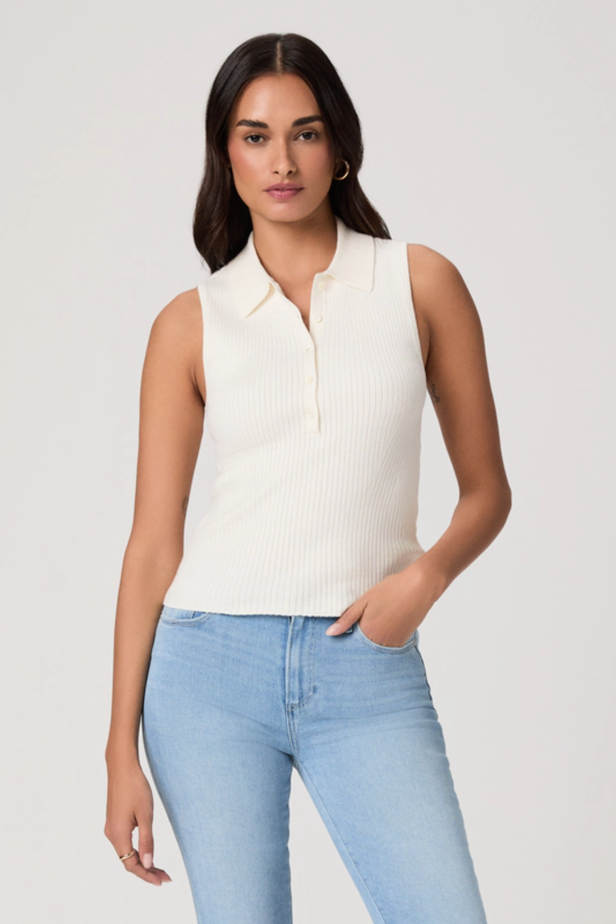 Woman wearing a white sleeveless sweater and light blue jeans against a plain background