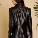 A woman shown from the back wearing a black faux-leather blazer with gold-tone buttons on the cuffs, paired with black pants, set against a warm beige background with decorative green leaves.