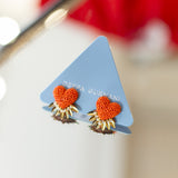 A pair of Deppa Gurnani earrings featuring textured orange-red hearts with gold sunburst details, displayed on a light blue triangular card.