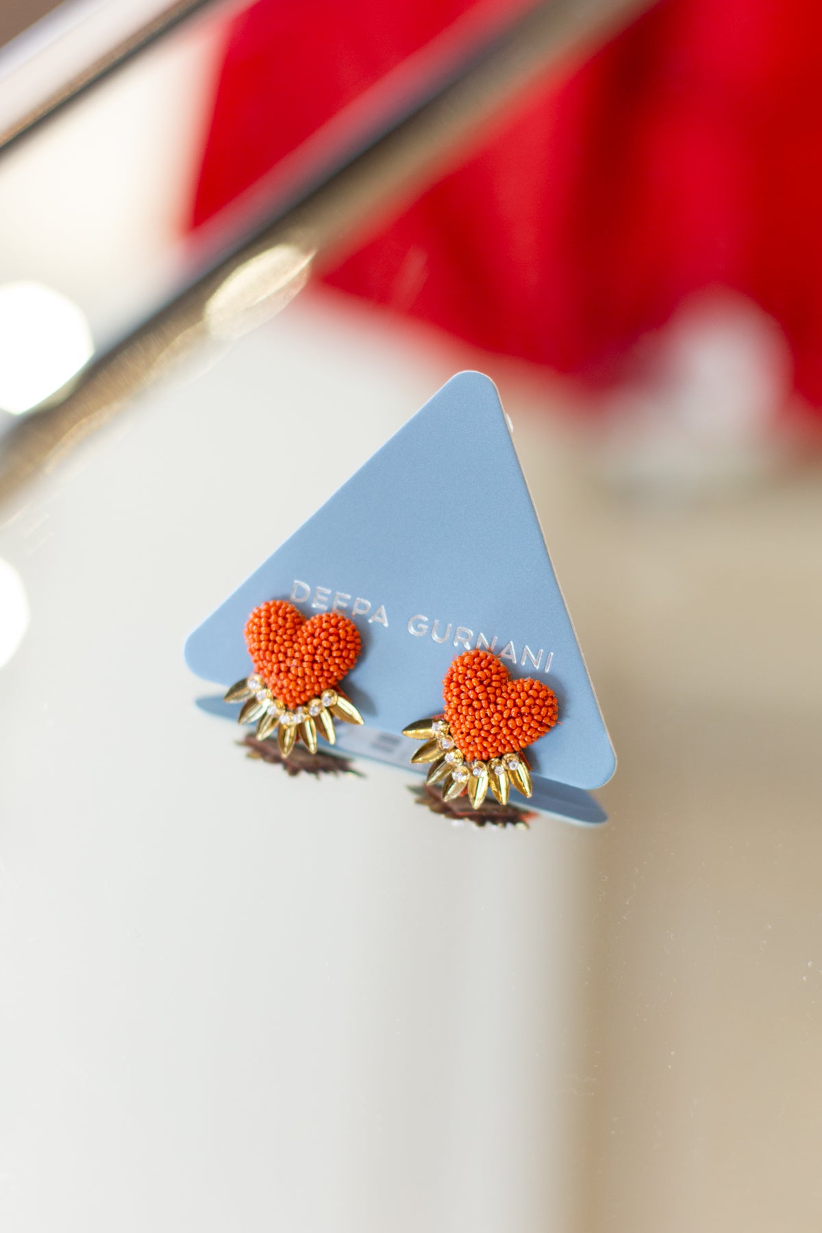A pair of Deppa Gurnani earrings featuring textured orange-red hearts with gold sunburst details, displayed on a light blue triangular card.