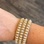 Three beaded stretch bracelets with white pearl-like beads and gold spacer beads, shown worn on a person's wrist against a blurred gray wood background.