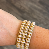 Three beaded stretch bracelets with white pearl-like beads and gold spacer beads, shown worn on a person's wrist against a blurred gray wood background.