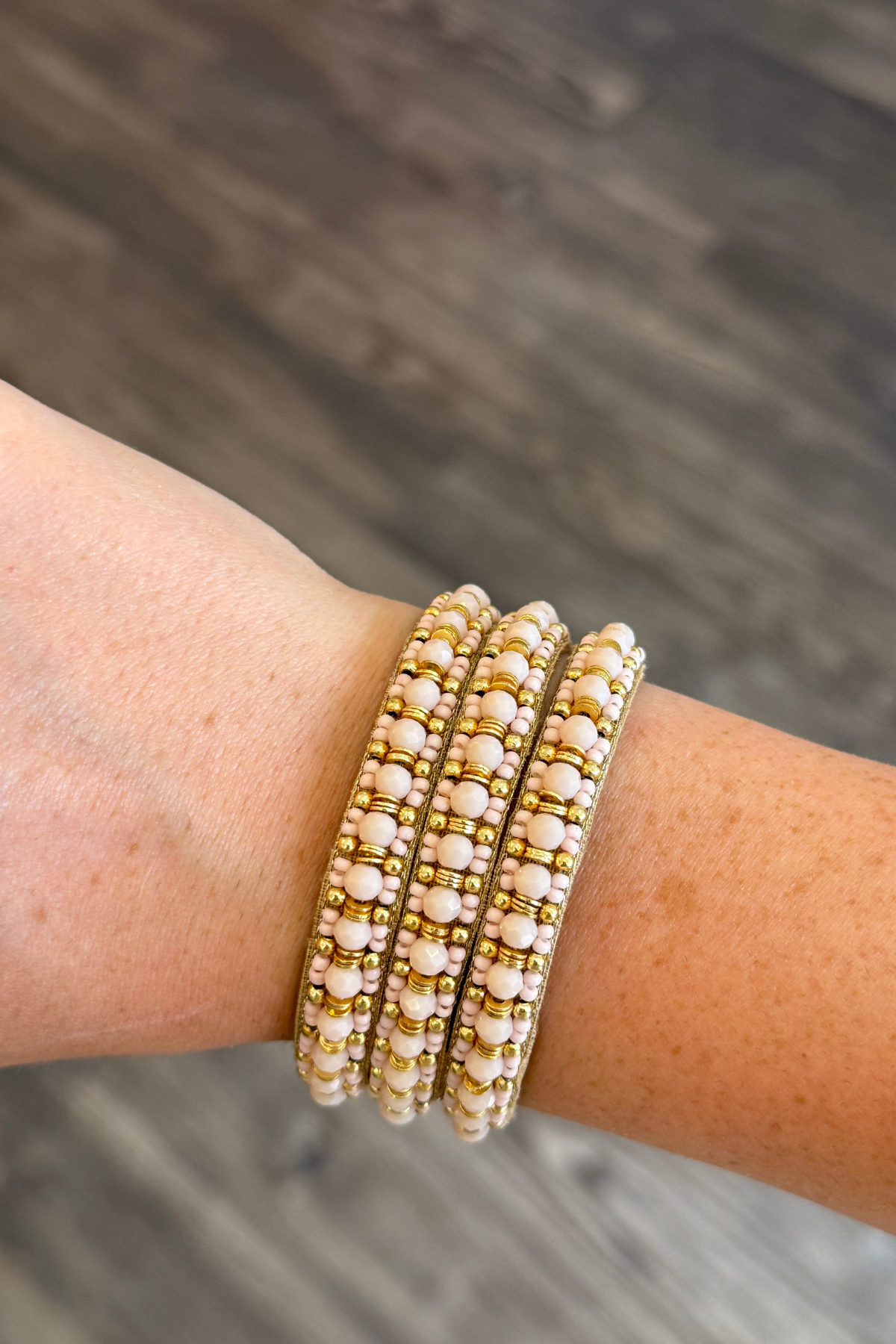 Three beaded stretch bracelets with white pearl-like beads and gold spacer beads, shown worn on a person's wrist against a blurred gray wood background.