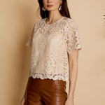 A woman modeling a short-sleeve cream lace top with a scalloped hem, paired with brown faux-leather pants, against a warm beige background.