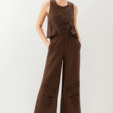 Woman wearing a brown sleeveless top and wide-leg pants with floral embroidery on a white background