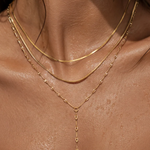 A close-up of a model's neck and upper chest wearing a layered gold necklace set, including a thin chain, a snake-style chain, and a delicate Y-shaped drop necklace.
