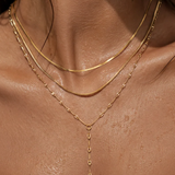 A close-up of a model's neck and upper chest wearing a layered gold necklace set, including a thin chain, a snake-style chain, and a delicate Y-shaped drop necklace.