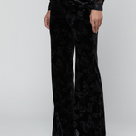 A model wearing black velvet wide-leg trousers with a subtle tonal butterfly pattern, paired with a sheer long-sleeve top and black pointed shoes, against a light gray studio background.