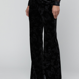 A model wearing black velvet wide-leg trousers with a subtle tonal butterfly pattern, paired with a sheer long-sleeve top and black pointed shoes, against a light gray studio background.