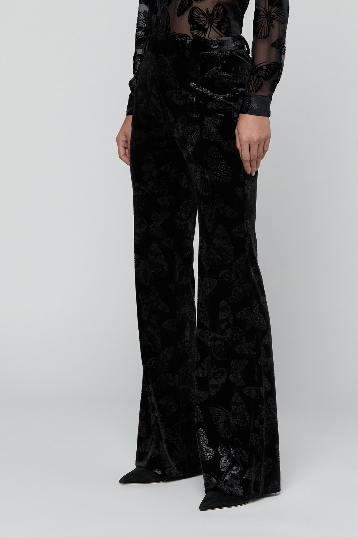 A model wearing black velvet wide-leg trousers with a subtle tonal butterfly pattern, paired with a sheer long-sleeve top and black pointed shoes, against a light gray studio background.
